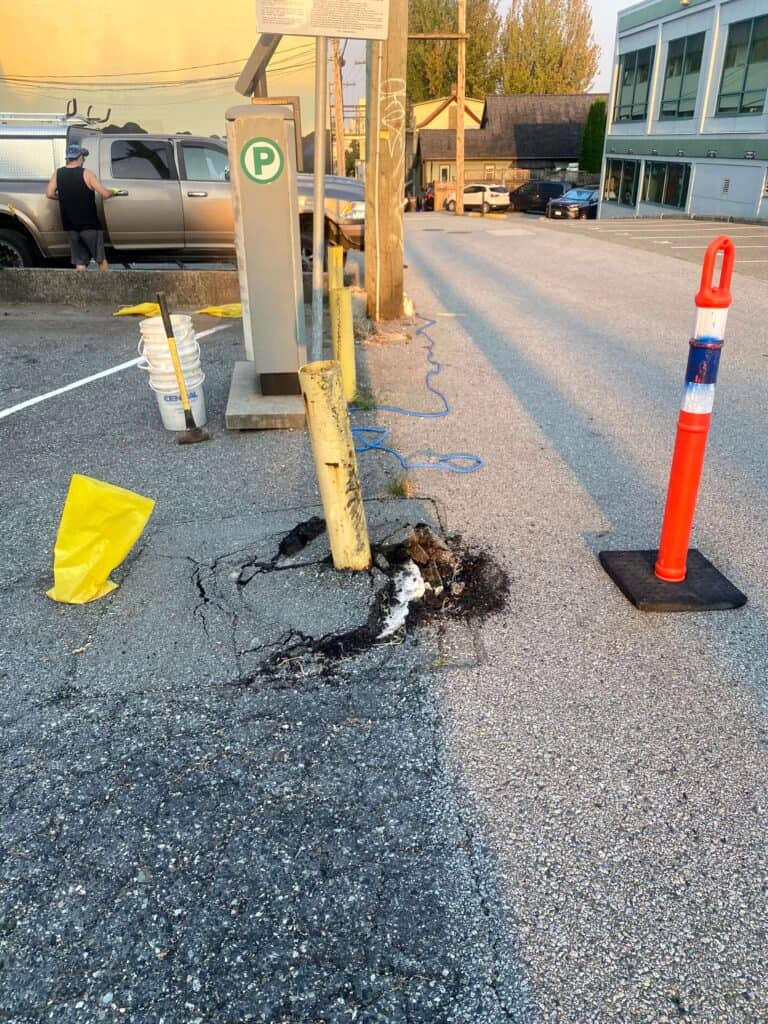A cracked concrete pavement in a parking lot features a damaged yellow post leaning to one side.