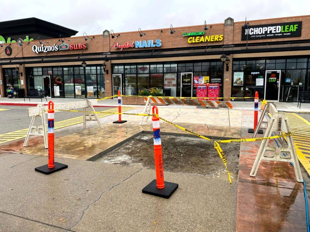A strip mall hosts a sandwich shop, nail salon, cleaners, and a restaurant. In the foreground, a construction zone displays freshly poured concrete cordoned off with caution tape and orange cones, covering part of the pavement.