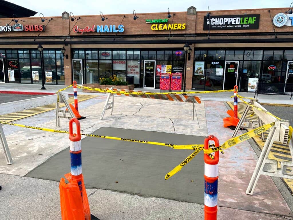A freshly poured concrete slab is cordoned off with orange and white traffic cones and yellow caution tape. The smooth, new surface lies in front of a row of shops, including a nail salon, cleaners, and a restaurant.