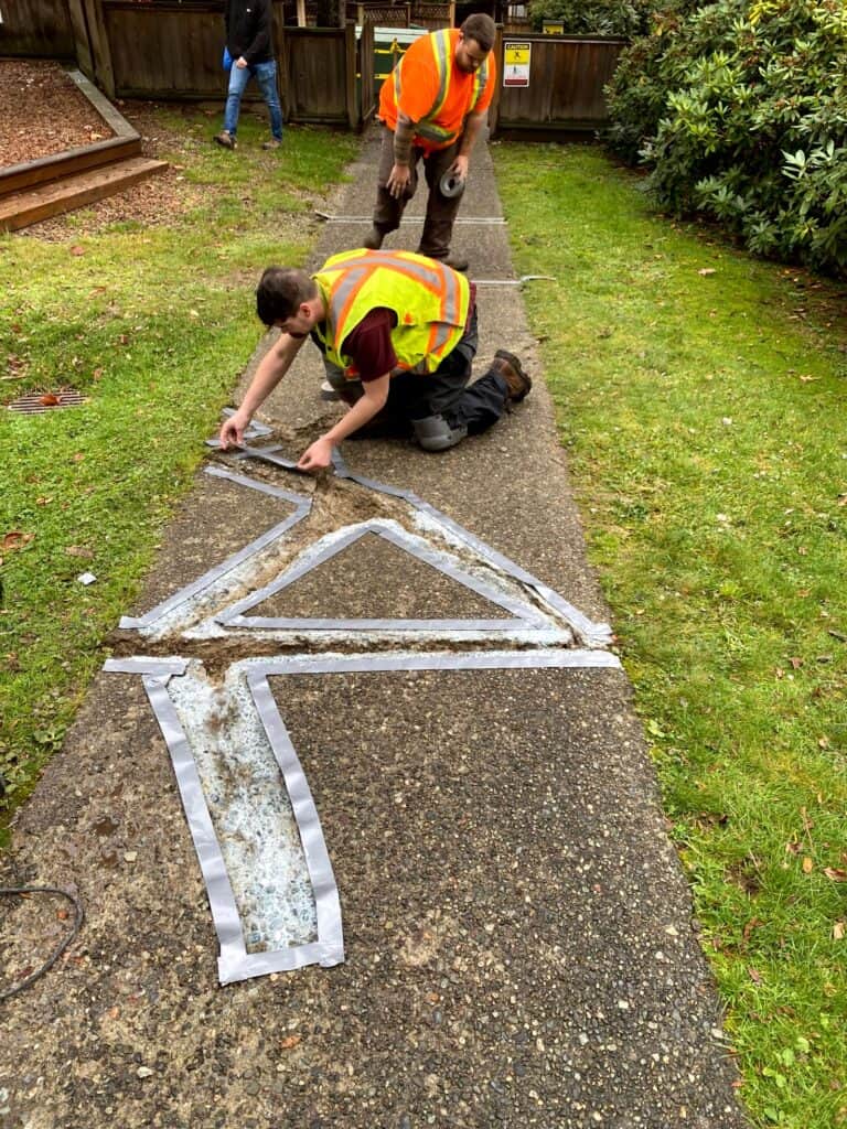 Two workers in safety vests diligently repair a cracked concrete path.