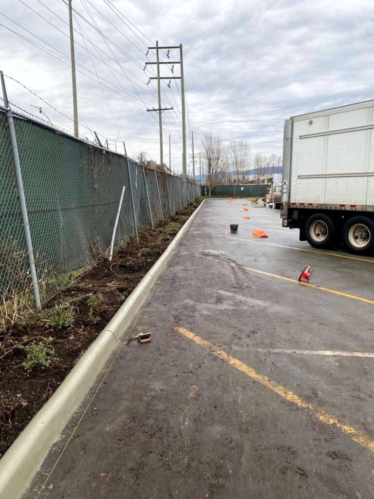A cleared, narrow dirt path runs along a fenced area beside a vast concrete parking lot.