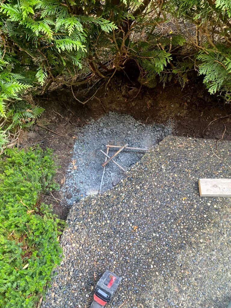 A section of the concrete path is broken, revealing exposed rebar underneath. Concrete surrounds green shrubs and soil. A piece of wood and a tape measure lie nearby on the concrete surface.